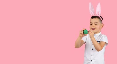 Beyaz tişört giymiş, elinde yeşil yumurta olan ve kafasında tavşan kulağı olan gülümseyen küçük çocuk, metni kopyalamak için fırçayla boyuyor..