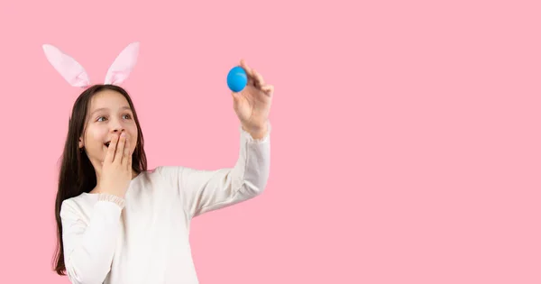 Дивовижна дівчина брюнетка покриває рот долонею і дивиться на пасхальне яйце блакитного кольору позує на рожевому фоні і копіює простір . — стокове фото