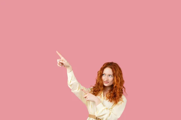 Рыжая девушка с длинными здоровыми и блестящими вьющимися волосами, указывая пальцами в сторону на копировальное место для рекламы на розовом фоне. — стоковое фото