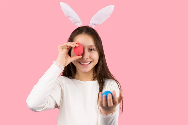 Närbild foto av en glad flicka med kaninöron på huvudet poserar med ett ägg håller i handen och med ett annat ägg täcker ett öga. — Stockfoto