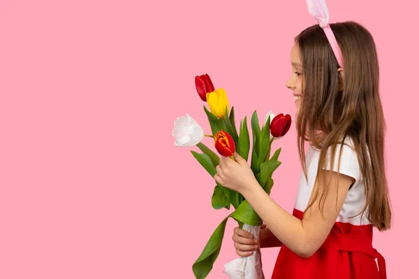 Vue de côté photo d'une jolie fille blonde aux cheveux longs portant des oreilles de lapin sur la tête et la robe tenant des fleurs dans ses mains sur fond rose et l'espace de copie. — Photo