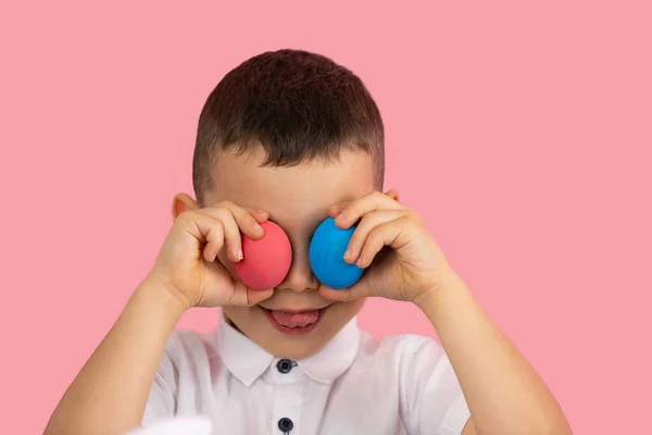 Niño en camiseta blanca cubriendo sus eyas con dos huevos de color rojo y azul y mostrando la lengua posando sobre fondo rosa. — Foto de Stock