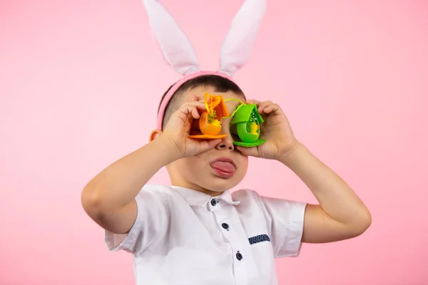 Närbild foto av en liten söt pojke bär kanin öron visar tunga och täcker sina ögon med leksaksägg på rosa bakgrund. — Stockfoto