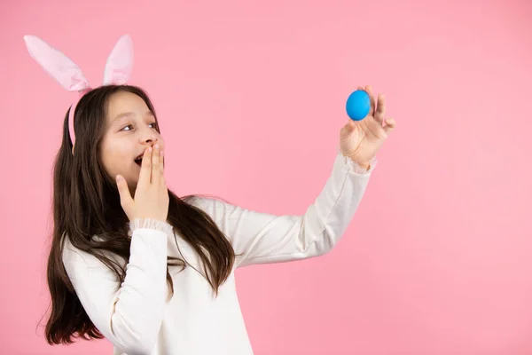 Sidovy foto av en brunett flicka med kanin öron på huvudet täcker hennes mun med handflata och tittar på blå ägg på utsträckt hand på rosa bakgrund. — Stockfoto