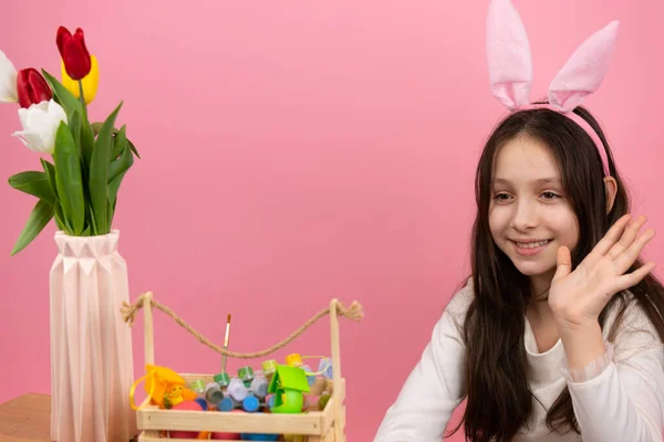 Söt liten flicka bär kanin öron på huvudet sitter vid bordet med en korg full av färger och vas med blommor viftar till kameran. — Stockfoto