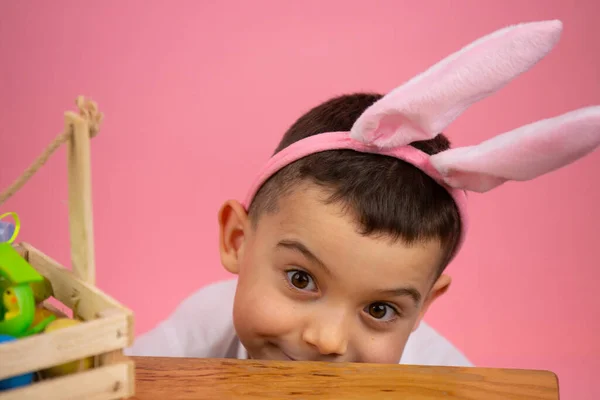 Joli garçon avec des oreilles de lapin sur la tête et les yeux largement ouverts posant sur fond rose avec un panier sur la table. — Photo