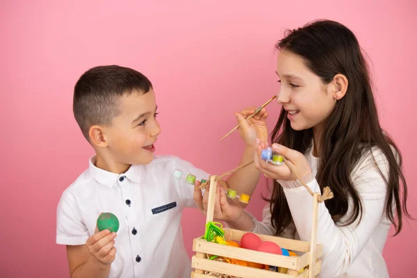 Lycklig familj förbereder sig för påsk, söt bror håller ett grönt ägg i handen och med hjälp av en pensel måla sin syster näsa medan hon håller en korg med ägg. — Stockfoto