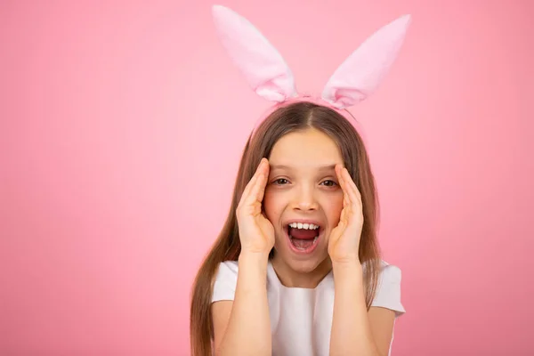 Närbild foto av en liten söt flicka bär kanin öron på huvudet tittar på kameran med öppen mun och täcker hennes ansikte på två sidor med handflatorna på rosa bakgrund. — Stockfoto