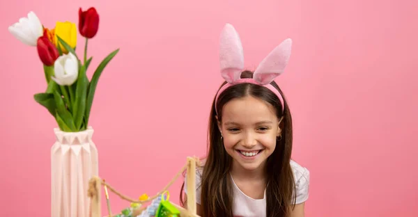 Närbild foto av en glad söt flicka med kanin öron på huvudet och en korg med färger på bordet och en vas med blommor, påsk koncept. — Stockfoto
