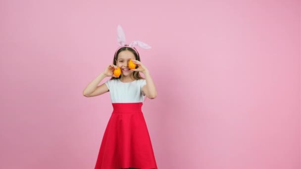 Video of a widely smiling girl with bunny ears holding two eggs of yellow color in her hands raises and lowers them in turn in front of her face. — Stock Video