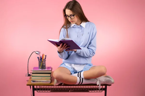 Fotografie vážné hezké školačky v uniformě s brýlemi čtení knihy připravující se na lekci sedí na stole se zkříženýma nohama. — Stock fotografie