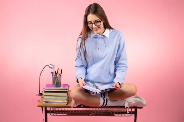 Mladý ležérní klidný dívka student v uniformě držení knihy a čtení sedí na stole se zkříženýma nohama v blízkosti stohu knih na růžovém pozadí. — Stock fotografie