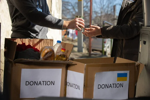 Cropped photo of a volunteer giving food to a refugee man standing near the car with many boxes of donation things inside the car. — Stock Photo, Image