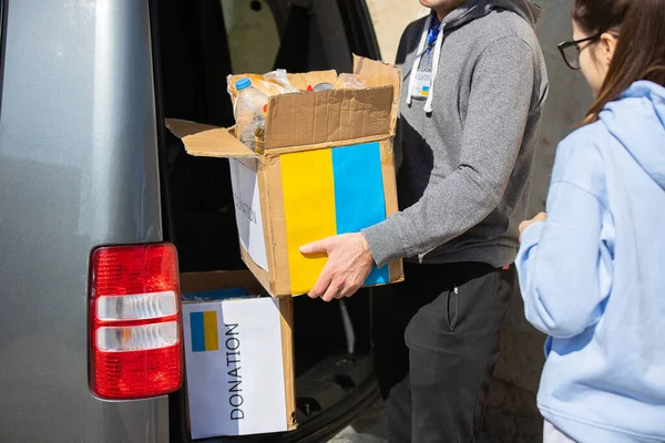 İki gönüllü genç kadın ve erkek Ukrayna 'dan gelen mültecilere yardım ediyor. Arabadan bir adam, Ukrayna bayrağında yiyecekle boyanmış bir kutuyu çıkarıyor..