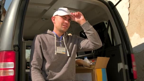 Vrijwilliger die op de kofferbak zit en zijn pet afdoet en zijn voorhoofd van de hitte veegt terwijl hij vluchtelingen uit Oekraïne helpt. — Stockvideo