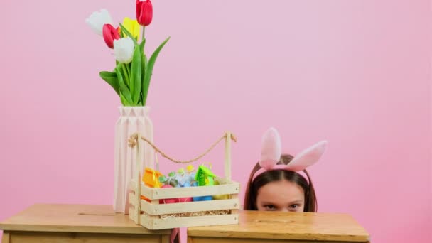 Vidéo d'une jolie fille souriante avec des oreilles de lapin sur la tête levant derrière la chaise avec panier et vase de fleurs il. — Video