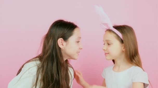 Twee vrolijke zussen, een van hen draagt konijnenoren op het hoofd, raken elkaar neuzen maken plezier op roze achtergrond. — Stockvideo
