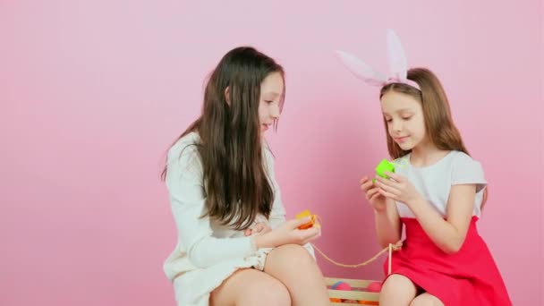 Twee mooie zusjes, een van hen droeg konijnenoren die hun cadeautjes wisselden terwijl ze samen zaten met een mand met beschilderd ei ertussen.. — Stockvideo