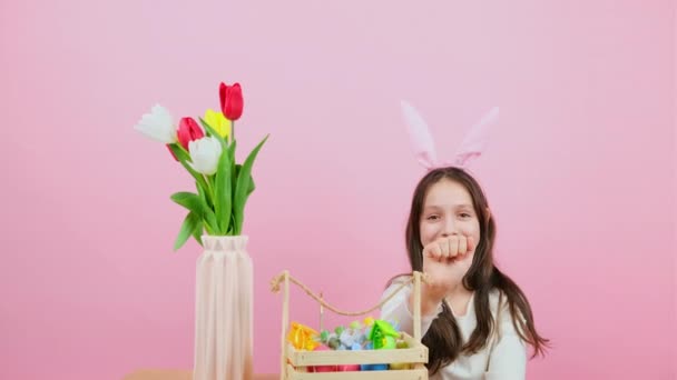 Glimlachend meisje met konijnenoren op het hoofd zittend achter twee stoelen met mand en vaas met bloemen die kloppen, wijzen en roepen gebaar. — Stockvideo