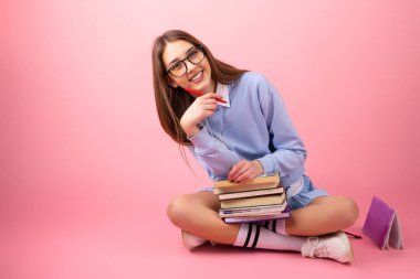 Gözlüklü neşeli liseli kız bacak bacak üstüne atmış, elinde bir yığın kitap ve pembe arka planda bir kalem tutuyor..