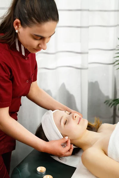 Foto completa de uma mulher massagista vestindo uniforme vermelho fazendo a uma mulher cliente massagens orelhas no salão de spa. — Fotografia de Stock