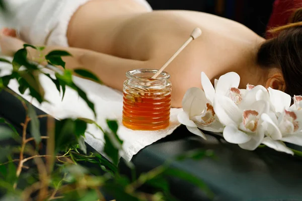 Young woman lying on the massage table with face down covered with towel and jar of honey near the woman for massage. — Stock Photo, Image