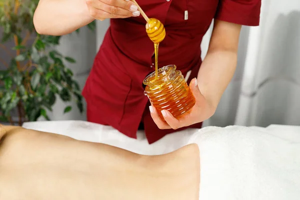 Foto de cerca de un frasco de miel que sostiene masajista en uniforme rojo para un procedimiento de masaje a una mujer joven acostada en la mesa en el salón de spa. —  Fotos de Stock