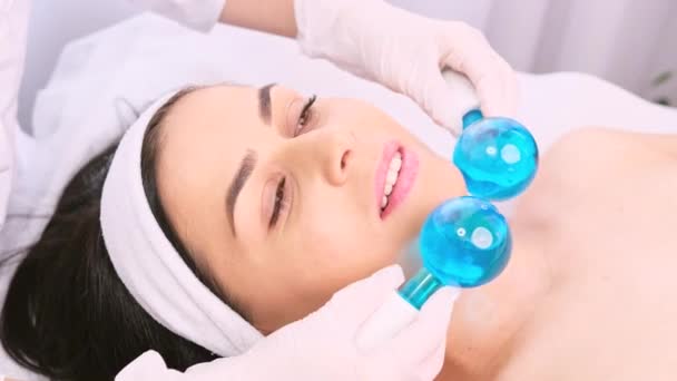 Hermosa mujer con diadema se somete a masajear el procedimiento facial por un cosmetólogo utilizando rodillo de hielo de bola azul en un salón de belleza. — Vídeo de stock