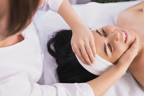 Above photo of cosmetologist making facial beauty massage treatment for relaxed young woman face with fingers movements in beauty spa salon. — Stock Photo, Image