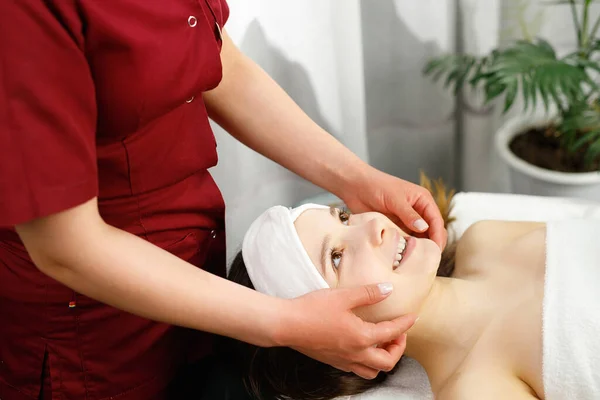 Giovane bella donna godendo procedura di massaggio viso da un massaggiatore specialista indossando uniforme rossa essere in salone spa. — Foto Stock