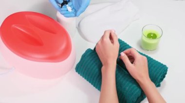 Koruyucu mavi eldivenli bir güzellik uzmanı krem şişesinden biraz krem alıyor ve güzellik salonundaki masada parafin banyosu ve mumla müşterinin ellerine masaj yapıyor..