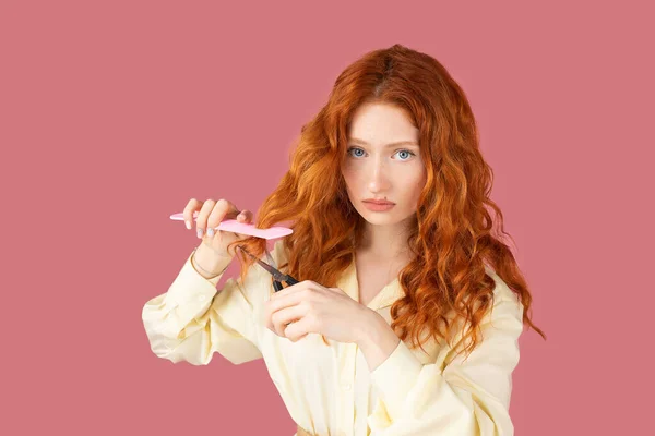 Menina ruiva bonita perturbada posando com tesoura e pente de cabelo mantendo em suas mãos e olhando para a câmera no fundo rosa. — Fotografia de Stock
