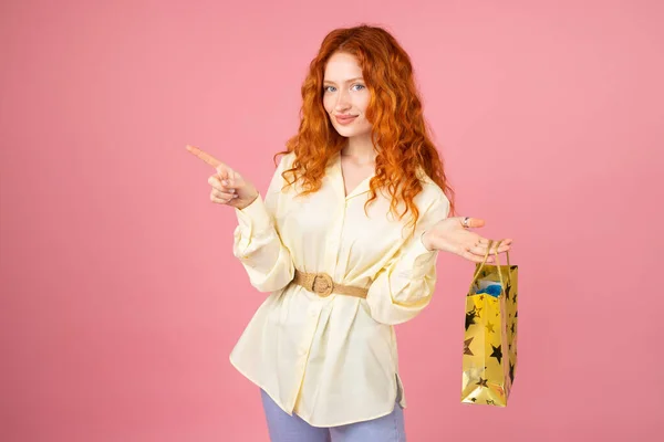 Glückliche rothaarige junge Frau hält in der einen Hand ein Paket mit einem Produkt für sich selbst und zeigt mit der anderen Hand zur Seite für Werbung. — Stockfoto
