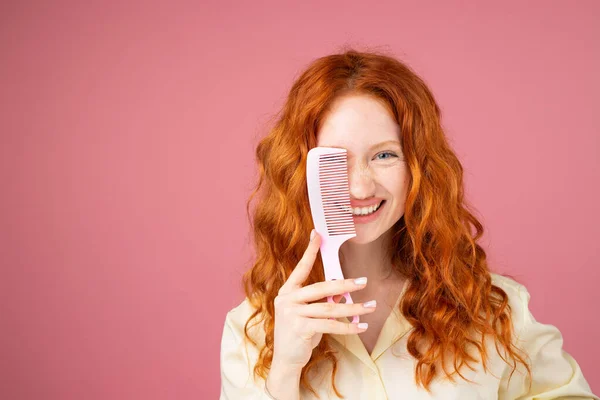Lächelnde junge Frau mit rotgelockten Haaren und blauen Augen posiert für die Kamera und bedeckt ihr ein Auge mit einem Haarkamm. — Stockfoto