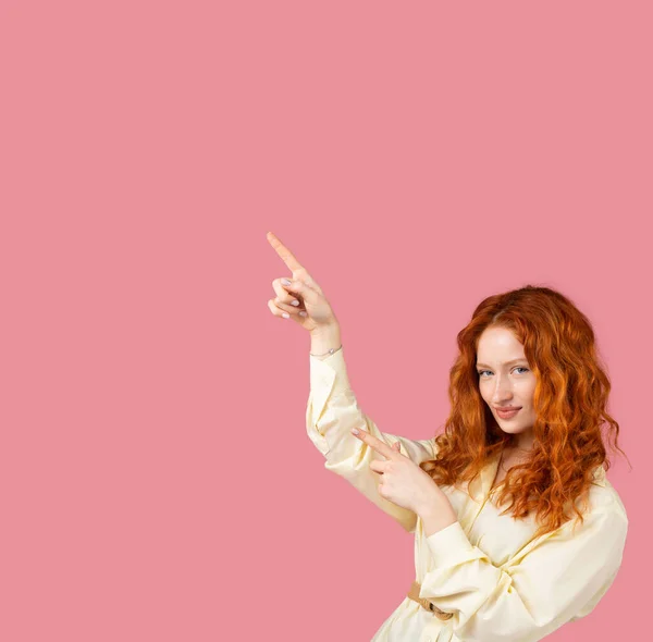 Привлекательная молодая женщина с рыжими волосами, смотрящая в камеру с тихой улыбкой и указывая двумя указательными пальцами на рекламу. — стоковое фото
