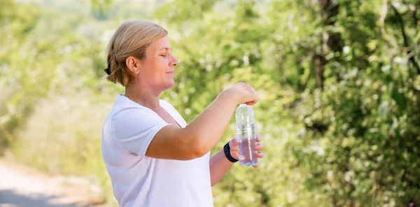 Photo vue de côté d'une femme blonde en plein air dans la forêt portant un t-shirt blanc et une montre intelligente essayant d'ouvrir la bouteille d'eau. — Photo