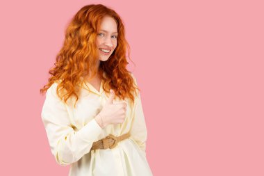 Gülümseyen kızıl saçlı genç kadın kameraya bakıyor. Pembe arka planda başparmağının görünmesi ve fotokopi çekmesi iyiye işaret..