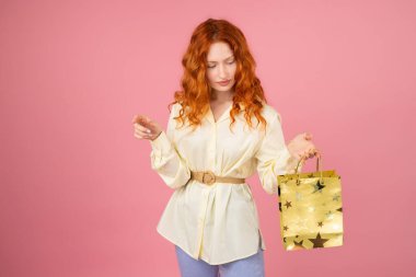Elinde sarı bir paket tutan, pembe arka planda poz veren güzel, genç bir kadın..