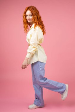 Full-length side shot of a beautiful woman with red hair standing on one leg and another raised back up on pink background.