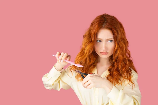 Portrait of a beautiful young woman with red hair and upset facial expression holding scissors and hair comb in her hands. — Fotografia de Stock