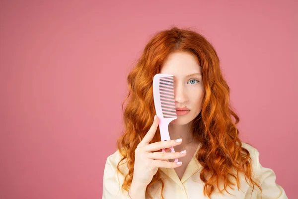 Foto de cerca de una pelirroja posando sobre un fondo rosado cubriendo un ojo azul con un peine de pelo y mirando fijamente a la cámara. —  Fotos de Stock