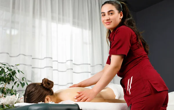 Foto completa del massaggiatore che guarda la fotocamera e fa la procedura di massaggio alla schiena a una giovane donna nel salone spa. — Foto Stock