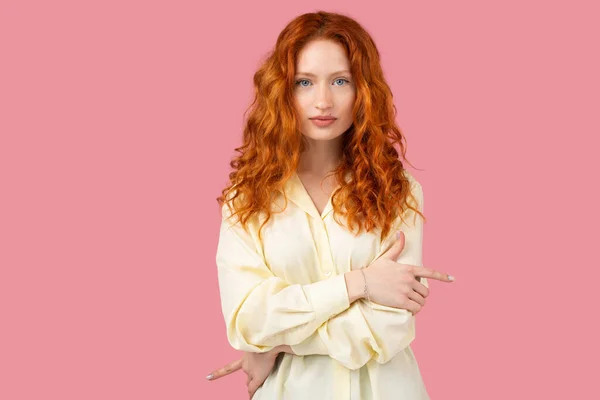 Beautiful elegant redhead freckled woman wearing yellow tunic folded her arms across her chest on pink background.
