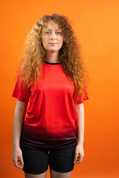 Redhead pretty woman football fan in red uniform of her favourite team posing for the camera on orange background. — Stockfoto
