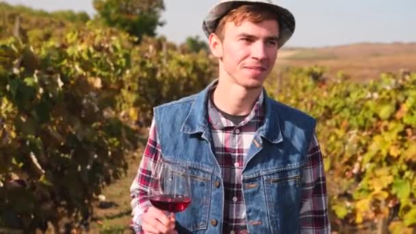 Close-up video van een gelukkige boer man in een hoed en denim vest lopen tussen de wijngaard met een glas wijn in zijn hand. — Stockvideo