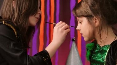 Little girl sitting without moving while another girl drawing a mask on her face for the Halloween party using brush and paints.
