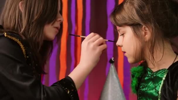Little girl sitting without moving while another girl drawing a mask on her face for the Halloween party using brush and paints. — Stok video