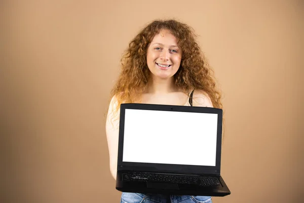 Young smiling woman with red curly long hair jolding in front of her opened laptop with white screen over golden background. — Foto Stock