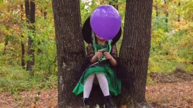 Ormanda iki ağacın arasında oturan üzgün bir kızın Cadılar Bayramı elbisesi ve elinde menekşe balonu tutan bir şapkayla videosunu kapat..