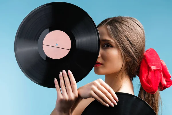 Side view photo of a gorgeous young lady with red ribbon on her hair tail keeping in the hands music records vynil over a blue background. High quality photo